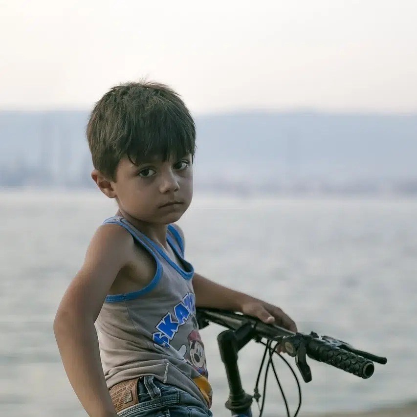 A young boy stands by the sea at dusk, holding onto a bicycle. His gaze is contemplative and a bit melancholic, suggesting a longing for support and care. This image captures the essence of the impact that donating to orphans in Islam can have, as it highlights the hopeful yet challenging circumstances faced by orphans awaiting kindness and generosity.