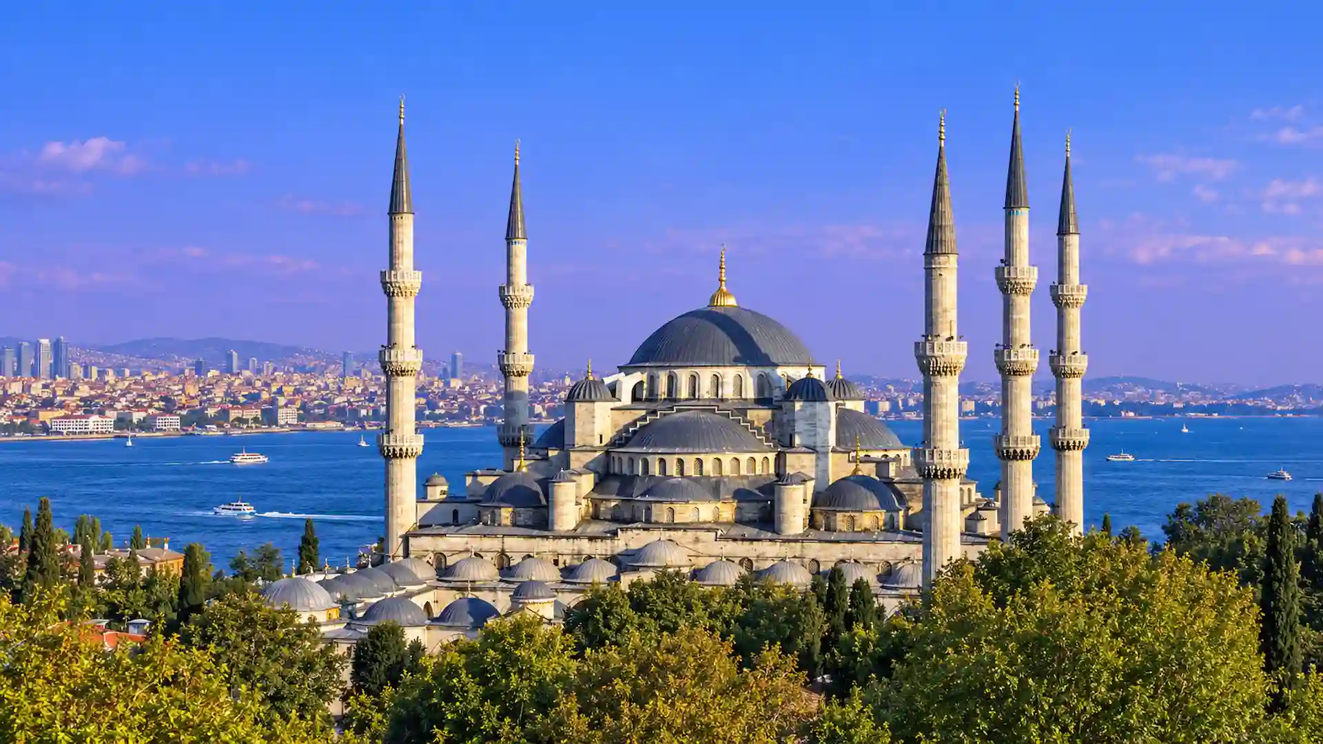 Panoramic view of the Blue Mosque in Istanbul, Turkey, featuring six towering minarets, cascading Ottoman domes, and the Bosphorus under a blue sky.