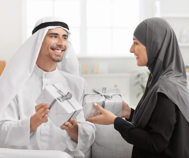 The image shows a happy moment between husband and wife, exchanging gifts. The man, wearing a white traditional Arabic thobe and a keffiyeh, smiles warmly as he holds a gift. The woman, wearing a dark-colored hijab and modest attire, smiles back at him while offering another gift.