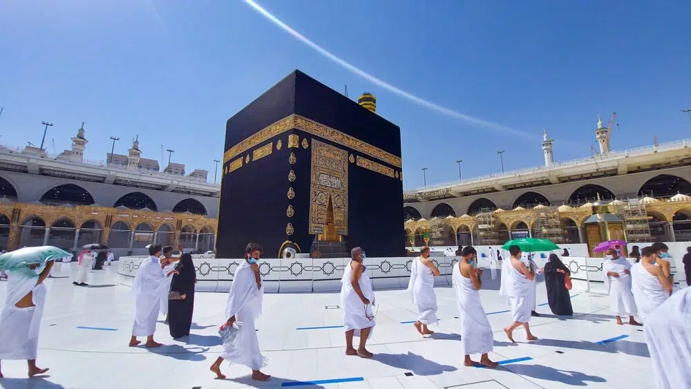 Pilgrims performing Tawaf around the Kaaba in Masjid al-Haram during Umrah, dressed in white Ihram garments under a clear blue sky.