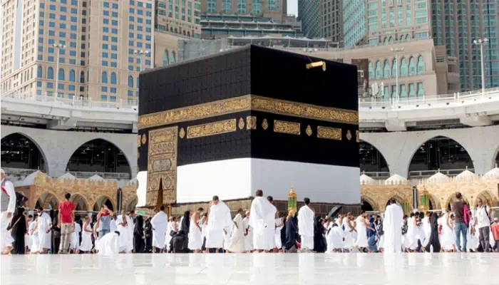 Pilgrims performing Tawaf around the Kaaba in Masjid al-Haram, Mecca, with tall buildings in the background, demonstrating the sacred ritual amidst a modern cityscape.