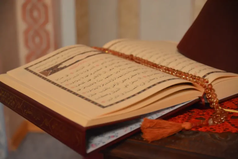 An open Quran placed on a wooden stand, with a prayer bead (tasbeeh) resting on it. The pages show Arabic text, beautifully illuminated, creating a serene and spiritual atmosphere.