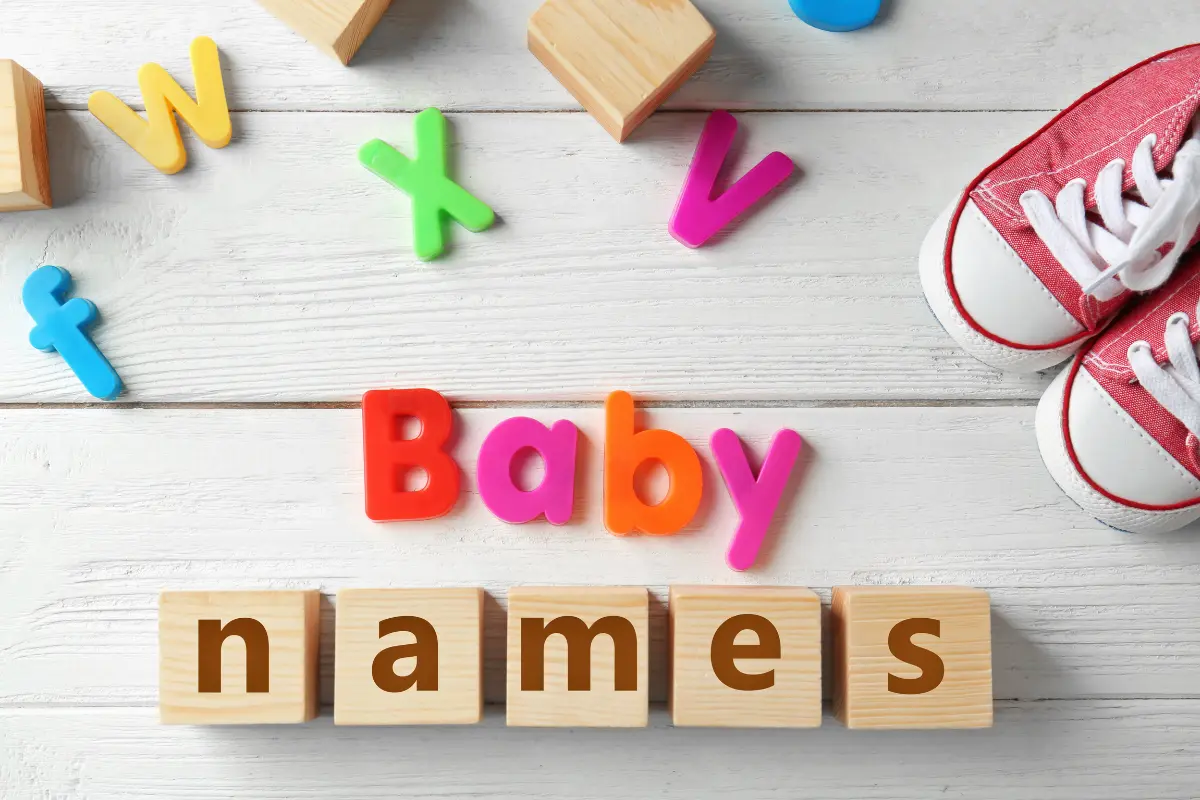 Colorful alphabet letters and wooden blocks spelling out 'Baby Names' on a white wooden surface, with red baby shoes placed on the side, symbolizing baby name selection.