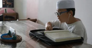 A young boy wearing a white cap and glasses studies the Quran online using a tablet, illustrating features to look for in a quality online Quran learning platform.