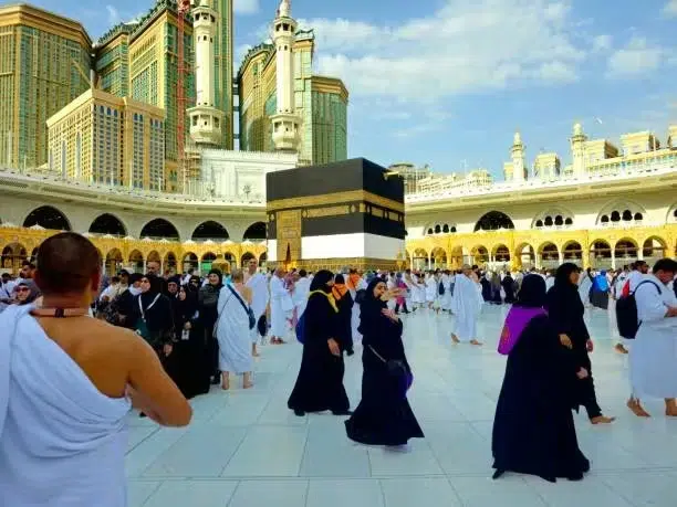 Muslim pilgrims performing Tawaf around the Kaaba at Masjid al-Haram in Mecca, with worshippers in white ihram and black abayas walking in the courtyard under a clear sky.